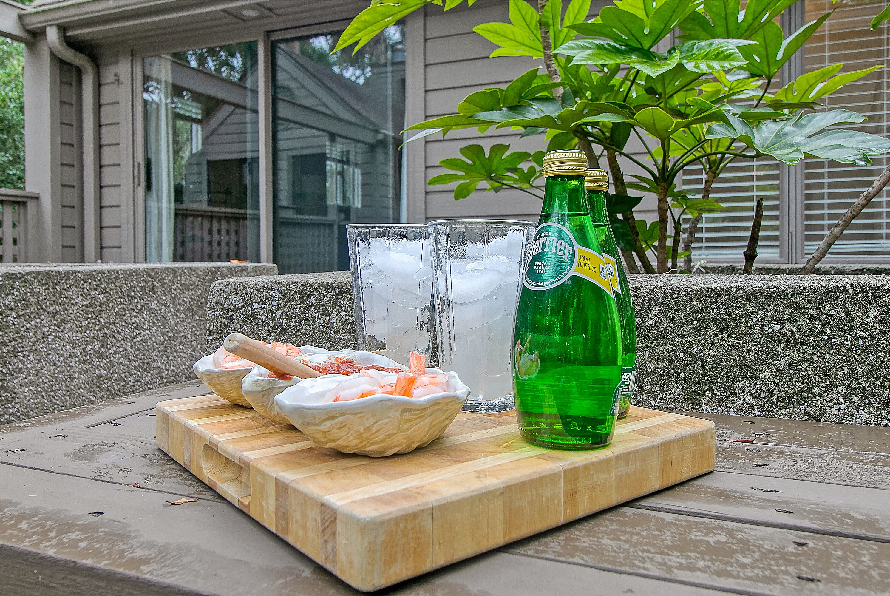 Een houten tafel met een groene fles, twee glazen met ijs en een schaal garnalen op een terras.