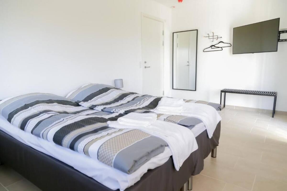 A neatly made bed with striped bedding, a bedside table, a wall-mounted TV, and a mirror.