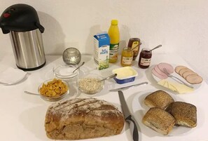 A table with a loaf of bread, cereal, butter, jam, a thermos, a carton of milk, a bottle of juice, and some sliced meat. - Hotel Ikast (Ikast)