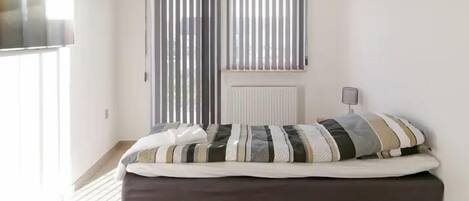 A bedroom with a bed, a radiator, a window with blinds, and a lamp on a bedside table.