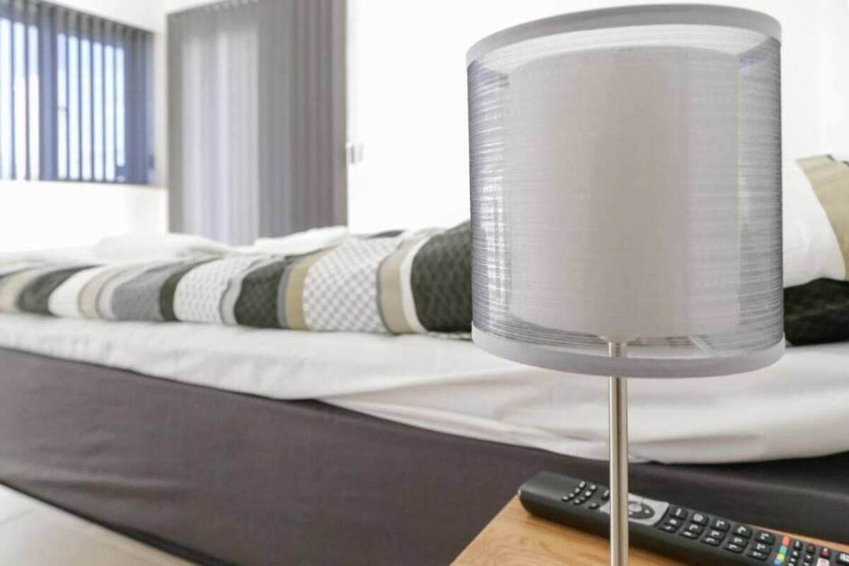 A bed with a grey headboard, a white blanket, and a striped pillow. A remote control is on a wooden surface in front of the bed.