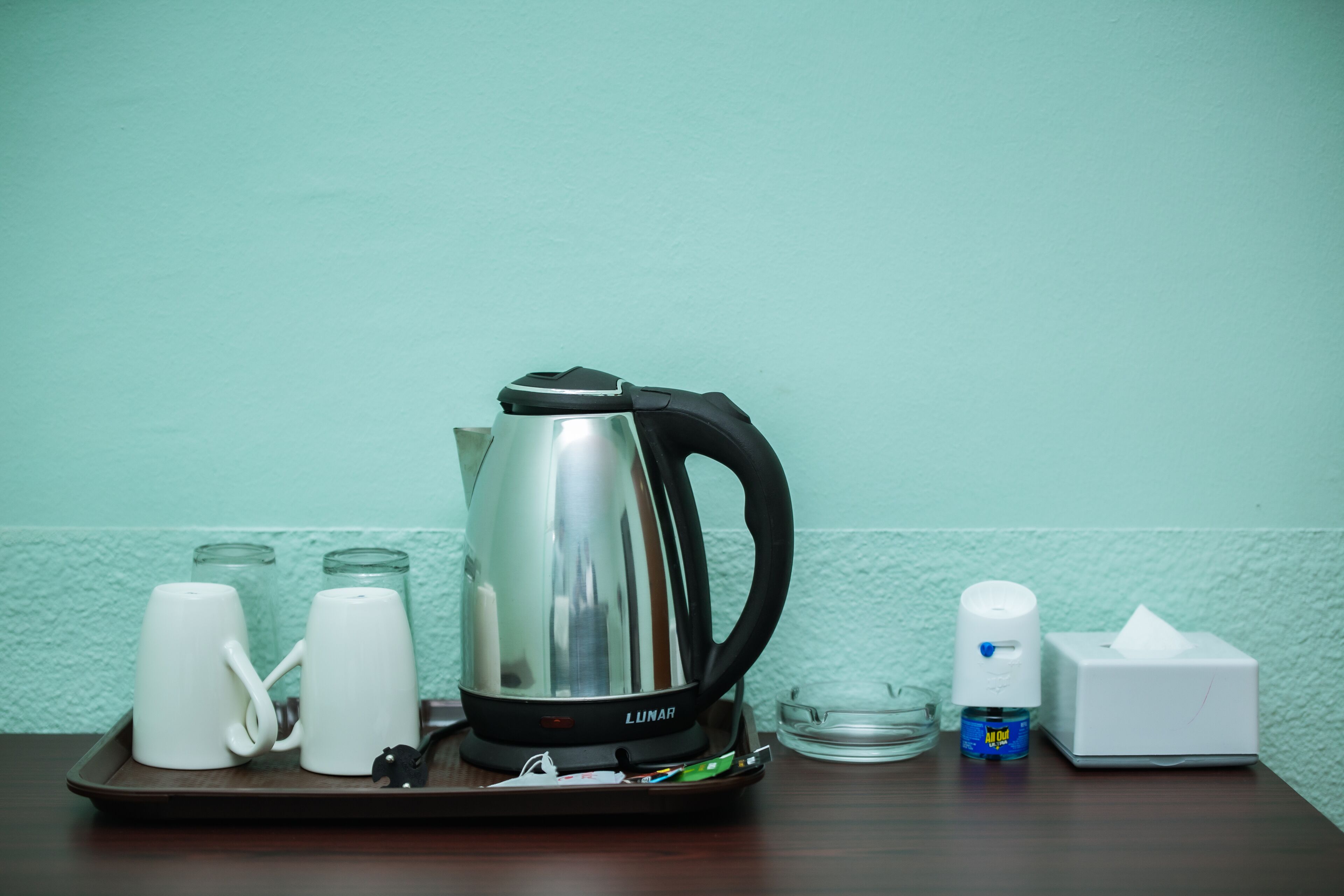 A kitchen area with a stainless steel kettle, two white mugs, a glass jar, a small bottle, and a tissue box on a tray.