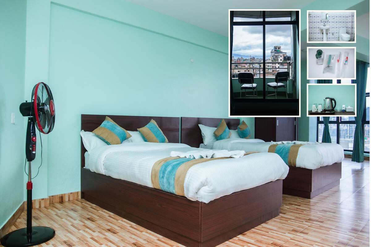 Habitación de hotel con dos camas, un ventilador y vista de la ciudad a través de amplias ventanas.