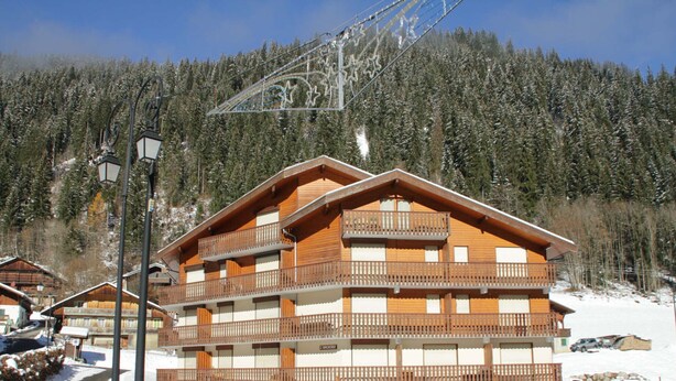 A multi-story wooden building with balconies, surrounded by snow and a forested mountain backdrop.