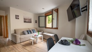 A modern living room with a sofa, a small table, and a dining table. There are two windows with curtains, a flat-screen TV mounted on the wall, and a door leading to another room. - Apartamentos Las Caldas . Castillo de Priorio , 15 Minutos del Centro de Oviedo (Priorio)