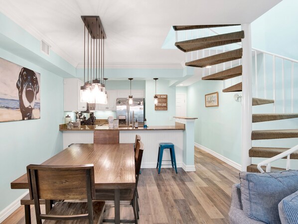 A modern kitchen with a central island, wooden dining table, and chairs. A staircase with a wooden railing leads to an upper level.