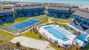 Un resort con varias edificaciones de techos azules, cancha de tenis, piscina y vista al mar.