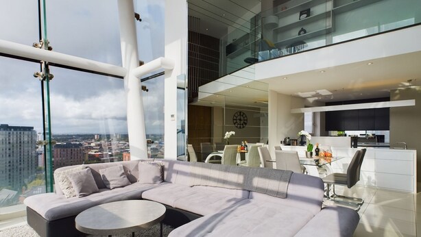 A modern living room with a large L-shaped sectional sofa, a round coffee table, and a glass dining table with chairs. The room features a large window with a city view, a kitchen area with modern appliances, and a glass railing overlooking the city. - Amazing Skyline Views 3BR Duplex (Manchester)