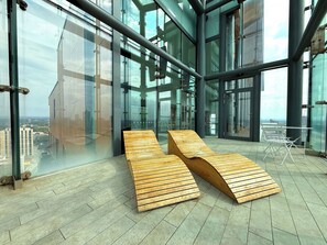 A modern interior with two wooden loungers, large glass windows, and a view of the cityscape. - Amazing Skyline Views 3BR Duplex (Manchester)