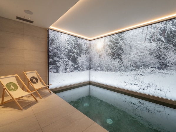 A modern indoor pool area with a large window displaying a snowy forest scene. There are two framed signs on the floor, one labeled "GENEPI LA PISTE" and the other "GRANDE PENTE 9".