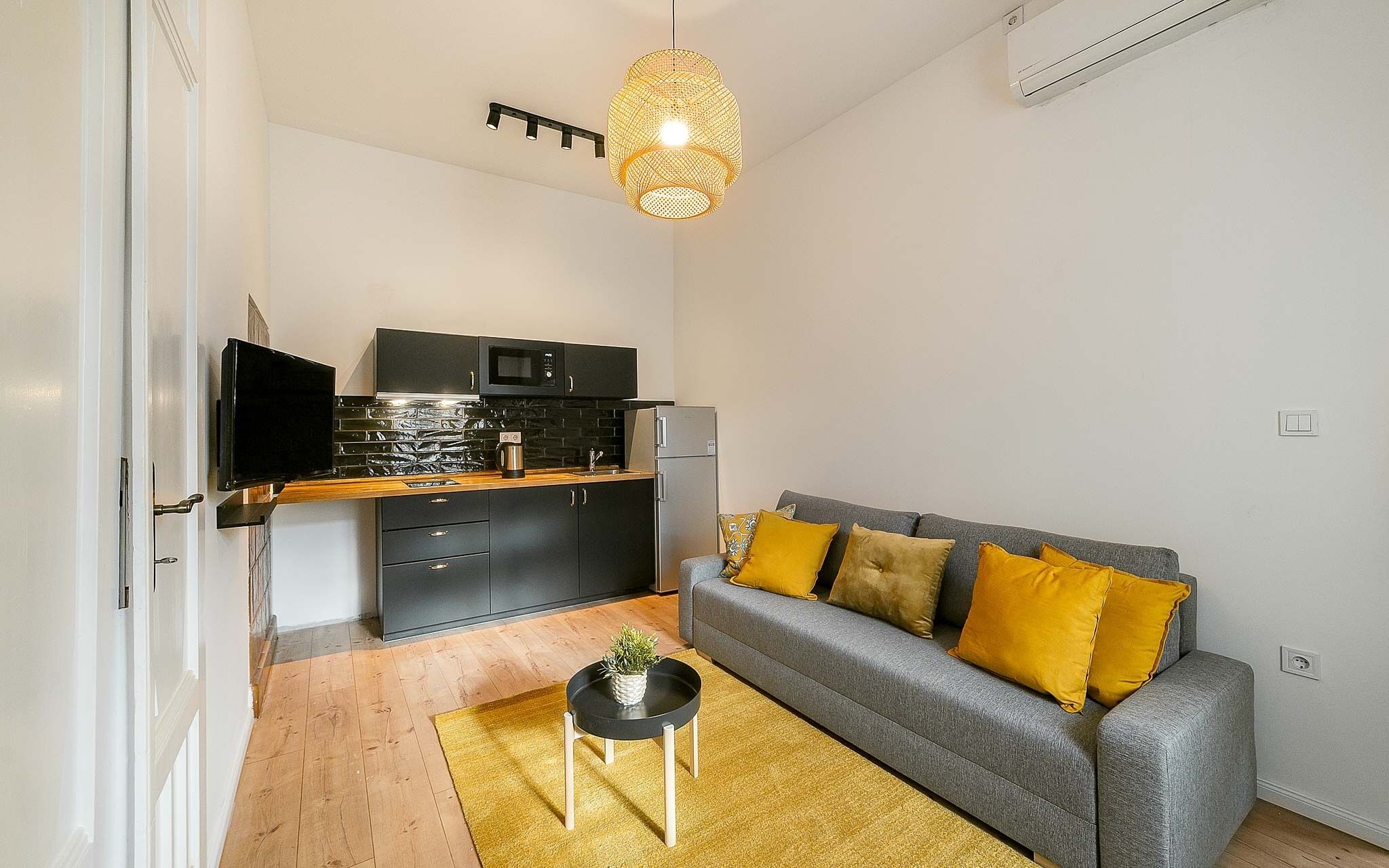 Un espacio habitable compacto con kitchenette, sofá gris con cojines amarillos, una pequeña mesita auxiliar y una lámpara colgante.