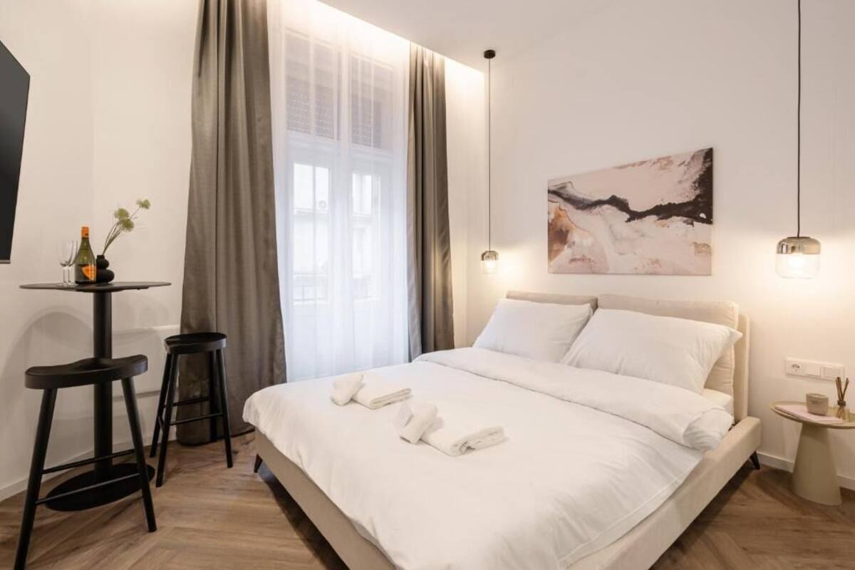Une chambre d’hôtel moderne avec un grand lit, une petite table sur laquelle se trouvent une bouteille et des verres, une chaise et un tableau accroché au mur.