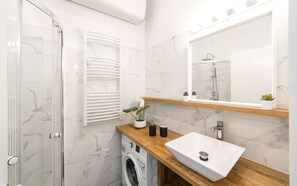 A modern bathroom with a glass shower enclosure, a built-in washing machine, a wooden vanity with a white sink, and a large mirror above the vanity. - A13 - Boutique Apartments by BQA (Budapest)