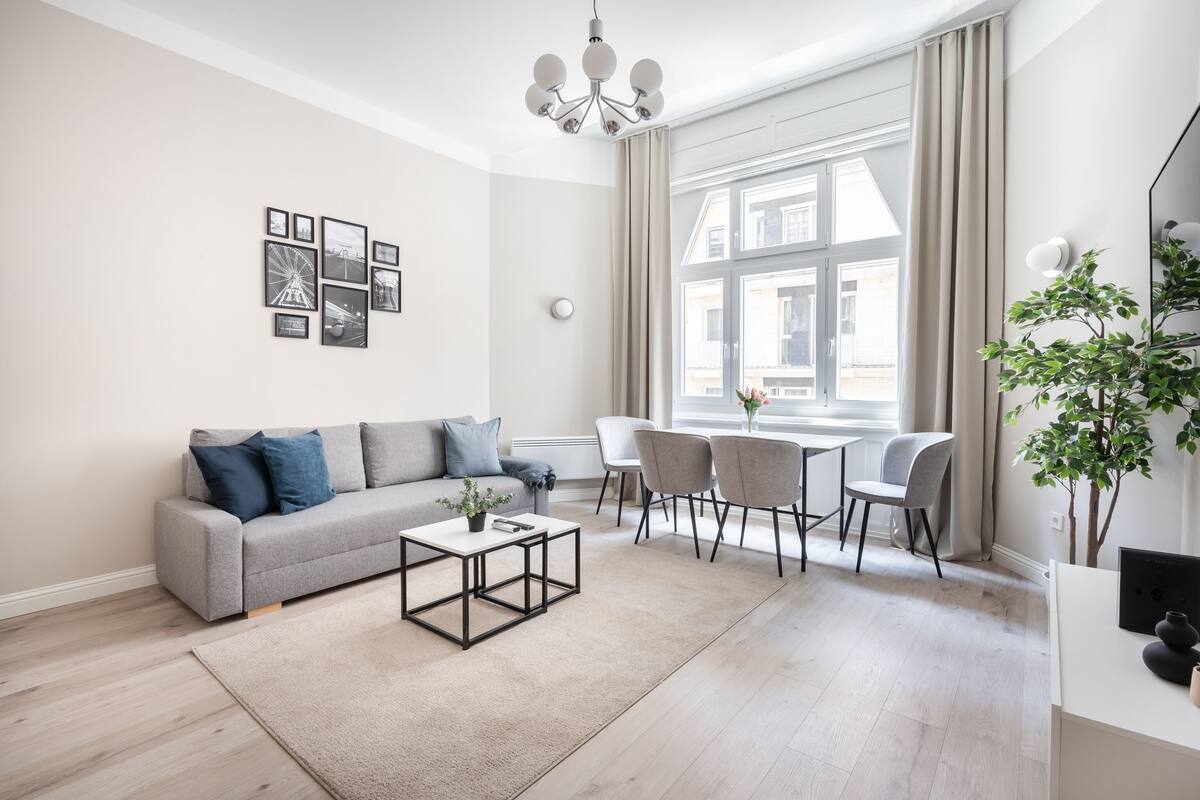 Un salon moderne avec un canapé gris, une table basse et un coin repas avec des chaises. Des tableaux encadrés ornent le mur, et une plante est placée à droite.