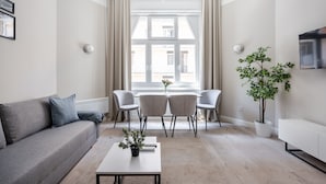Una sala de estar moderna con un sofá, una mesa de centro con una planta, una zona de comedor con sillas, un televisor y un ventanal grande con cortinas.