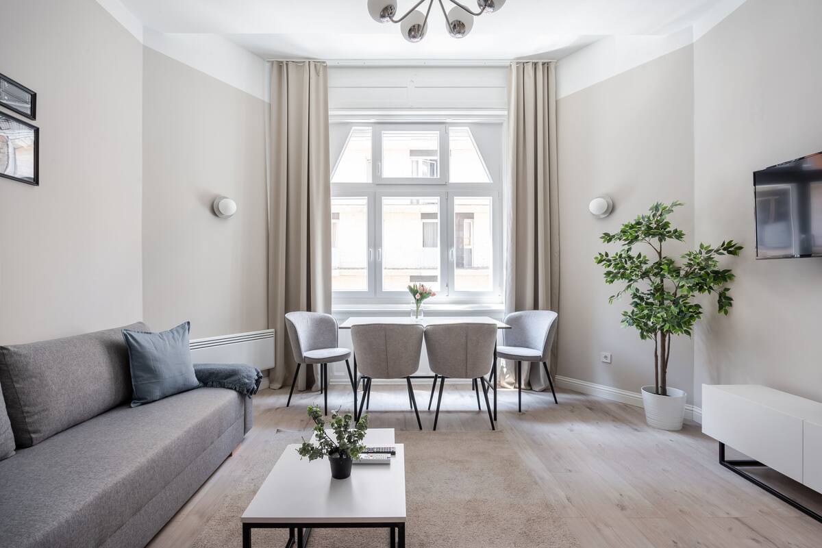 Un salon moderne comprenant un canapé, une table basse avec une plante, un coin repas avec des chaises, un téléviseur et une grande fenêtre avec des rideaux.