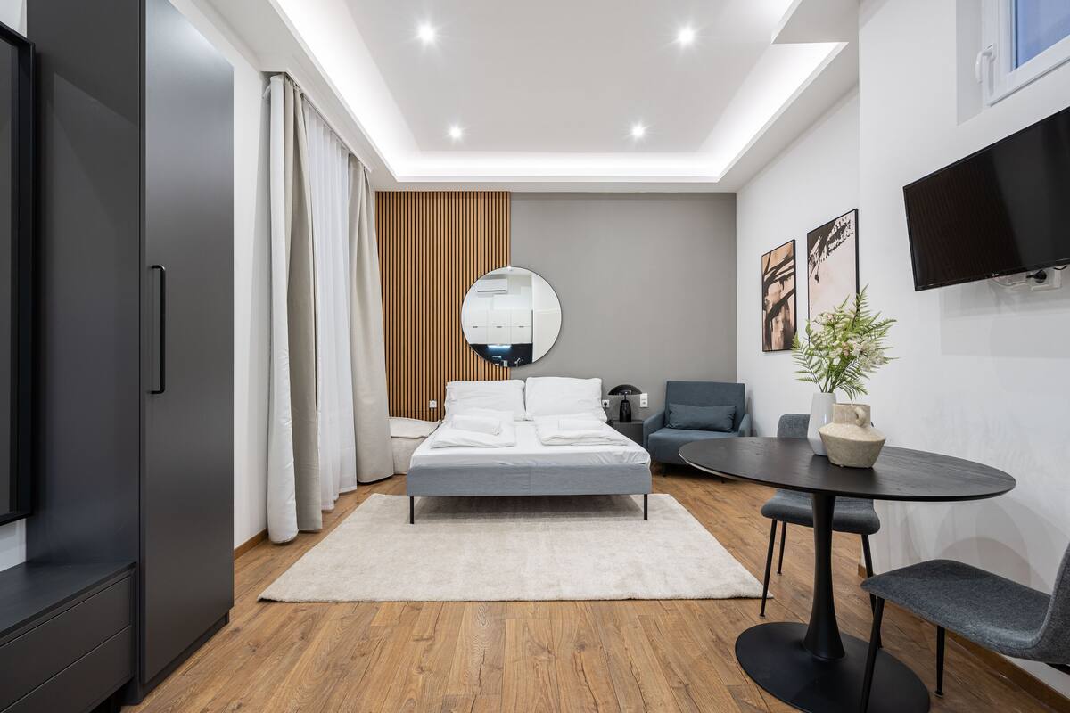 Une chambre d’hôtel moderne avec un lit, un miroir rond, une télévision, une table à manger et une plante.