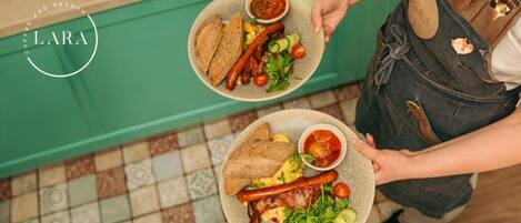 A person holding two plates of food, one with sausages and salad, and the other with eggs and bacon, in a cafe setting.