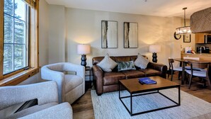 A living room with a sofa, armchairs, a coffee table, and a dining area with a table and chairs. There are two wall-mounted lamps and a ceiling light. - Corner Unit w/Mountain Views, River Run Village (Keystone)