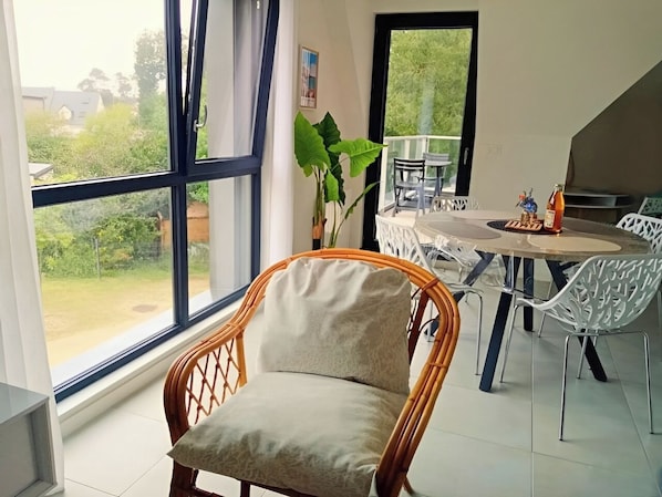 Une pièce dotée d'une grande fenêtre, d'un fauteuil en osier, d'une table à manger avec des chaises et d'une plante en pot.