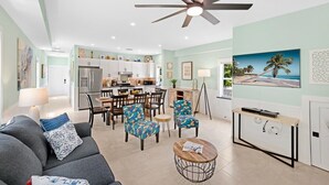 A modern living room with a sofa, chairs, a coffee table, and a TV stand. There is a dining area with a table and chairs. The room has a ceiling fan and a kitchen in the background.