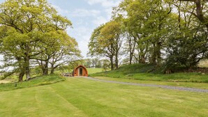 A rustic wooden structure with a rounded archway, surrounded by lush green grass and trees. - Conwy Pod (Llanrwst)