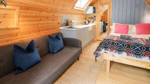 A cozy cabin interior with a wooden ceiling, a sofa with blue cushions, a bed with a patterned blanket, and a kitchenette area. - Conwy Pod (Llanrwst)