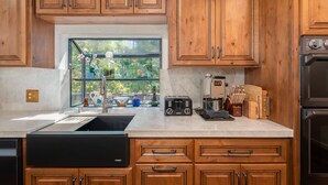 A kitchen with wooden cabinets, a window with plants, a sink, a toaster, and a coffee maker. - Huckleberry House- Rec Room (Arnold)