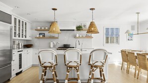 Cocina moderna con armarios blancos, una isla central con taburetes y lámparas colgantes de mimbre, además de una zona de comedor con sillas de madera.