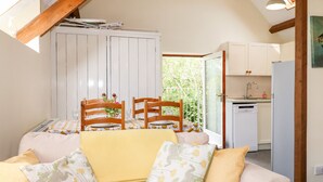 A cozy living room with a sofa, a dining table with chairs, a kitchen area with cabinets, and a window overlooking greenery. - Higher Norton Barn (Totnes)