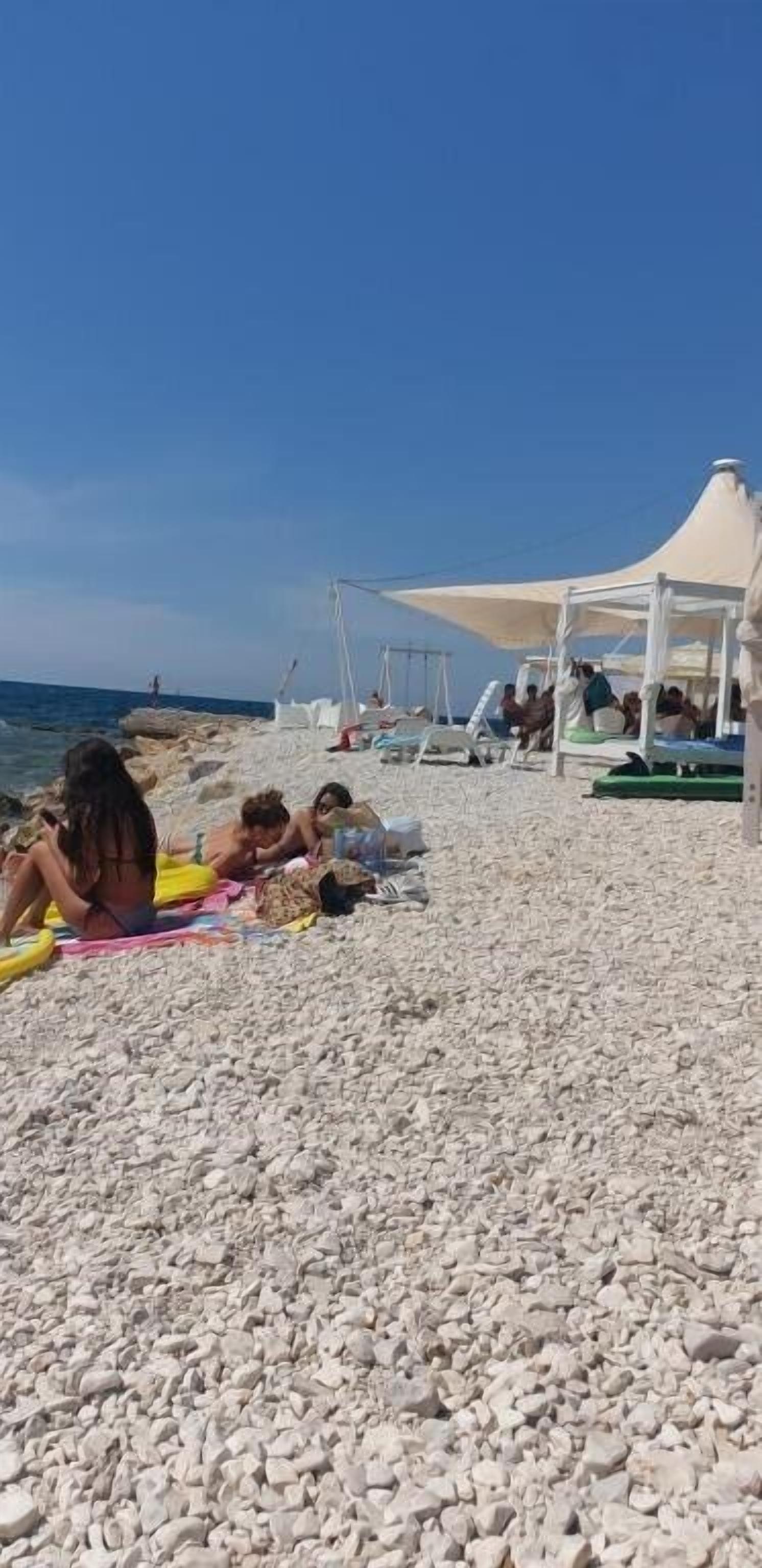 Una spiaggia con ciottoli, persone che prendono il sole e una struttura a tenda bianca.