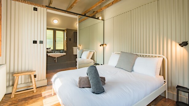 Une chambre à coucher avec un grand lit, des poutres en bois, un petit tabouret, un miroir et une salle de bain visible à travers une porte ouverte.