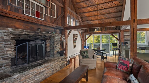 L’intérieur d’un chalet rustique, avec une cheminée en pierre, des poutres en bois, un coin salon confortable et une vue sur l’extérieur.