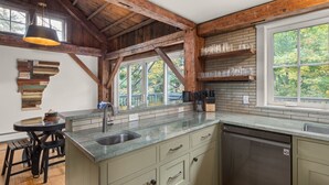 Une cuisine dotée d’un plan de travail en pierre, d’un évier et d’un lave-vaisselle. Le plafond est en bois et une fenêtre offre une vue sur les arbres.