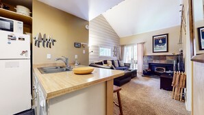 A compact kitchen with a sink, a microwave, and a refrigerator. There is a dining area with a sofa and a fireplace. - La Residence V #t05 1 Bedroom Condo by RedAwning (Mammoth Lakes)