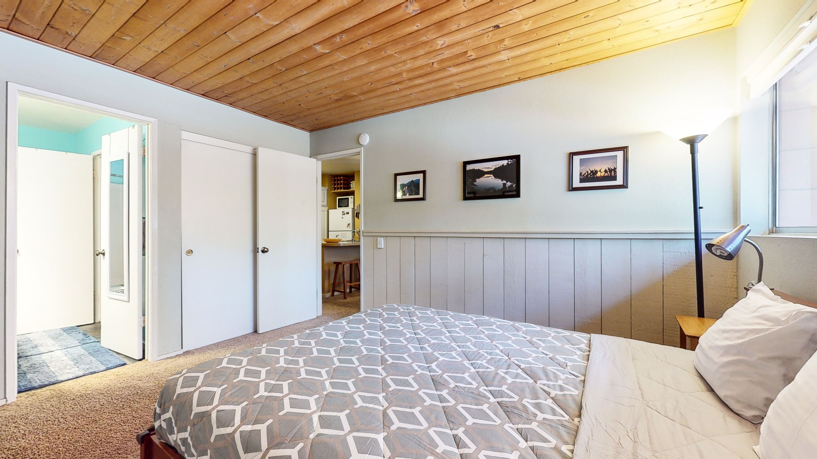 A bedroom with a wooden ceiling, a bed with a patterned bedspread, a nightstand with a lamp, and a wall with framed pictures.