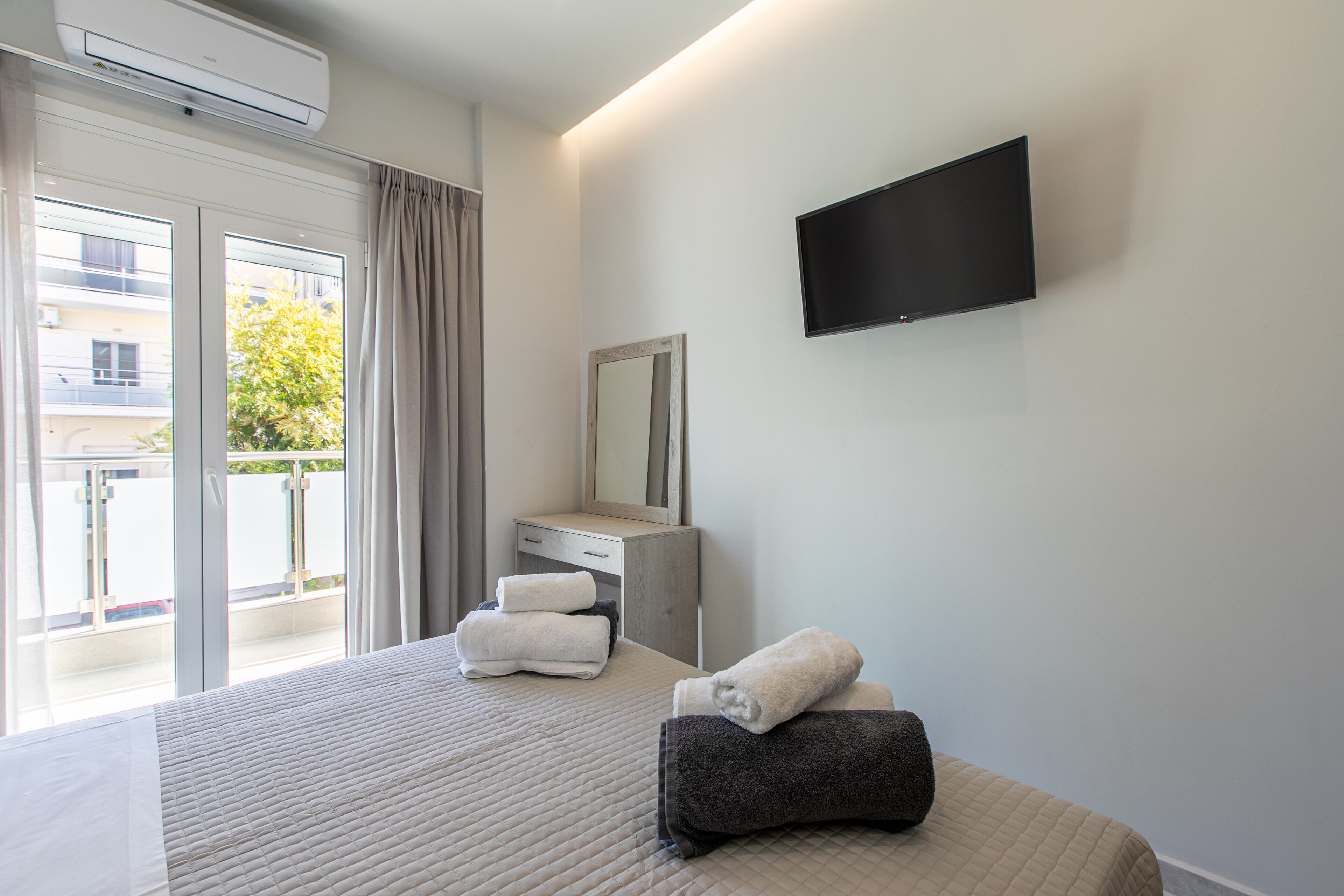A hotel room with a bed, a flat-screen TV, a dresser, a mirror, and a balcony with curtains.