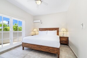 A bedroom with a wooden bed, two bedside tables with lamps, an air conditioning unit, and a large window with a view of greenery.