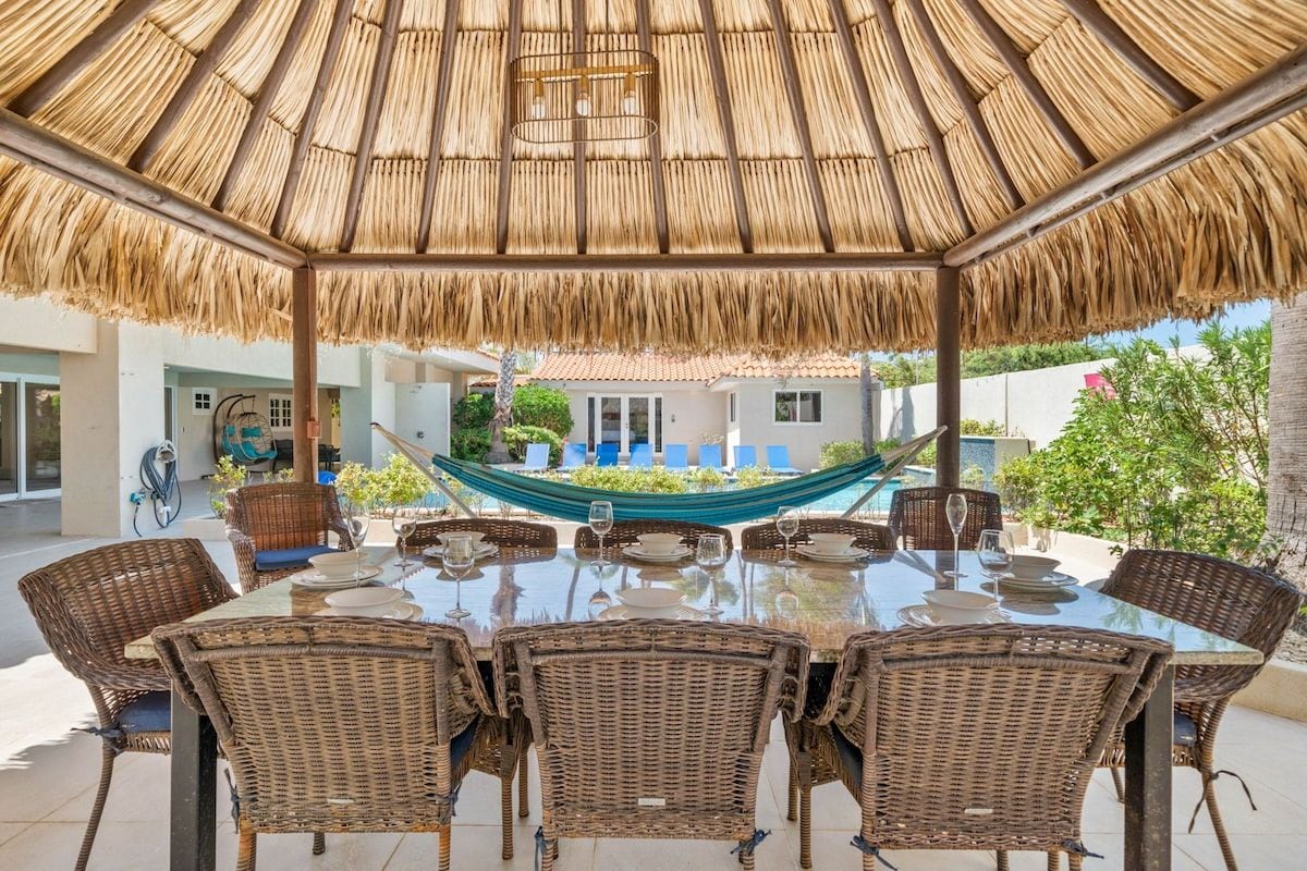 Een terras met een rieten dak, rieten meubels en een uitzicht op een huis op de achtergrond.