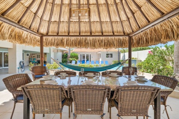 An outdoor dining area with a thatched roof, wicker furniture, and a view of a house in the background.