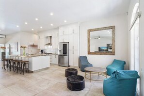 A modern kitchen with white cabinets, a central island, and bar stools. There are two bean bags and a small round table.