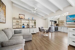 Ein modernes Wohnzimmer mit einem Ecksofa, einem Essbereich mit zwei Stühlen, einer Küche inklusive großem Fernseher und einem Deckenventilator.