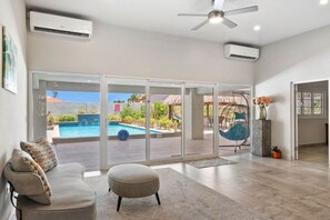 A spacious living area with a sofa, ottoman, and a view of a swimming pool and outdoor seating.