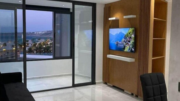 A modern living room with a large TV, a black sofa, and a view of a cityscape through floor-to-ceiling windows. - Appartement de Luxe Avec vue sur mer à Malabata (Tanger)