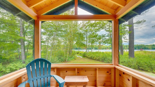 A wooden cabin with a large window offering a view of a forest and lake. - Dock & Beach Access! Waterfront Home on Kezar Lake (Lovell)