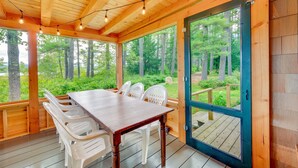 A wooden deck with a table and chairs, large windows, and a view of the forest. - Dock & Beach Access! Waterfront Home on Kezar Lake (Lovell)
