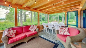 A wooden deck with wicker furniture, string lights, and large windows overlooking a forest. - Dock & Beach Access! Waterfront Home on Kezar Lake (Lovell)