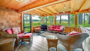 A wooden interior with wicker furniture, a fireplace, and large windows offering a view of the outdoors. - Dock & Beach Access! Waterfront Home on Kezar Lake (Lovell)