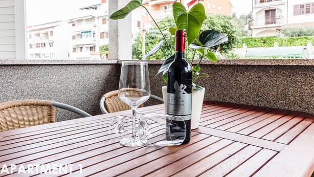 A table with a bottle of wine, a glass, and a plant on a balcony with a view of buildings. - A cozy apartment, 1 km from the beach in a quiet neighborhood (Vrsar)