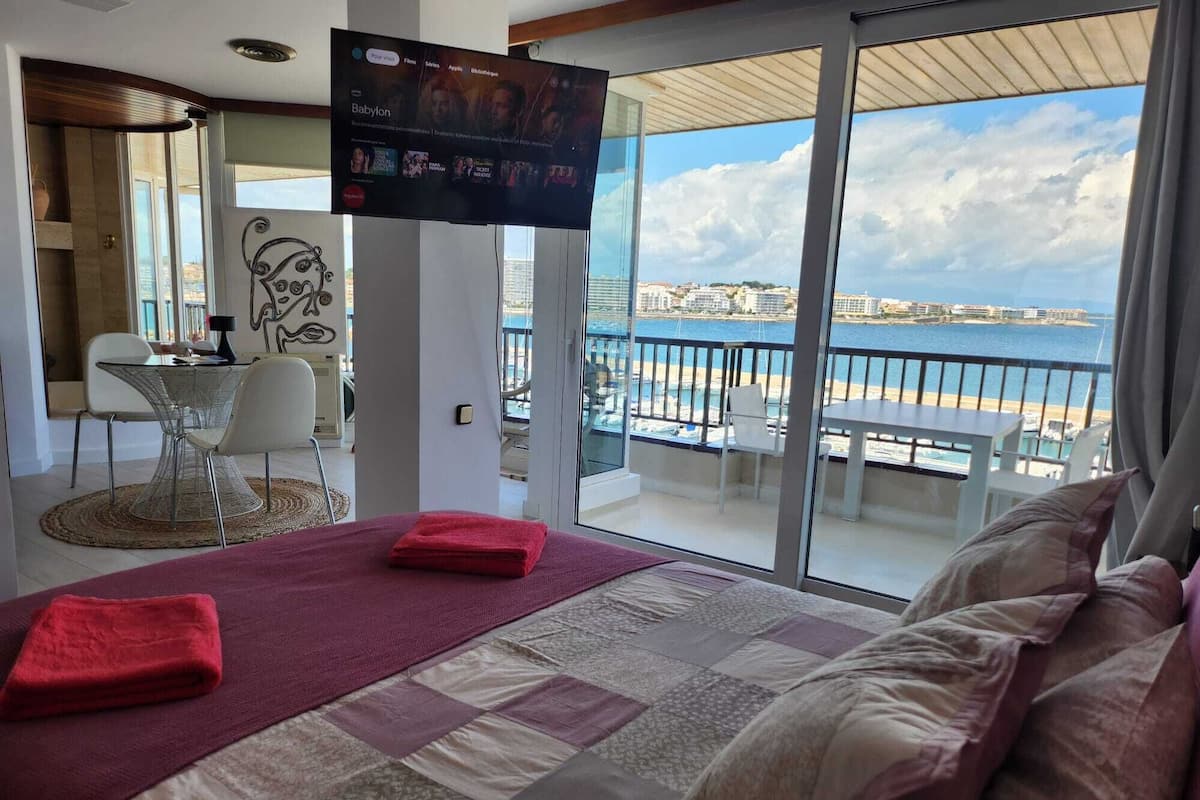 A hotel room with a large balcony, a bed with a red towel, a TV on the wall, and a view of the sea and buildings.